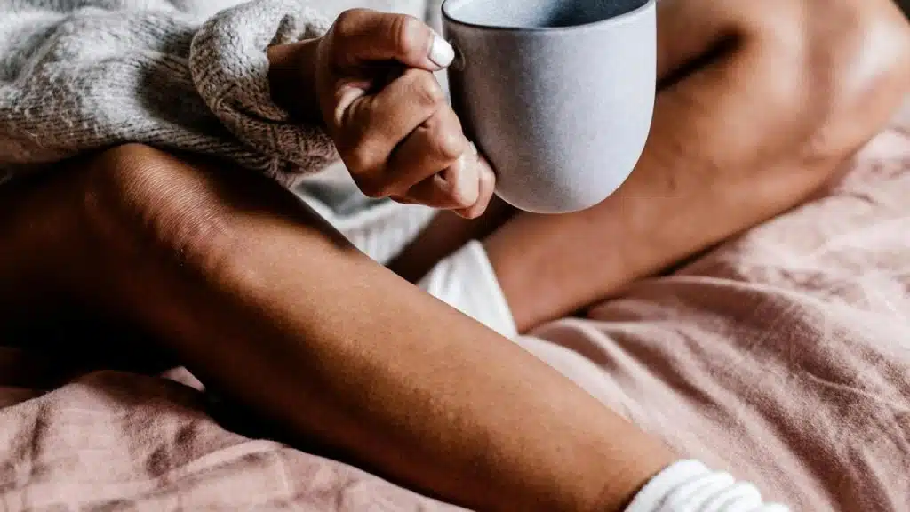 A person sitting on a bed holding a mug while wearing a cozy sweater.