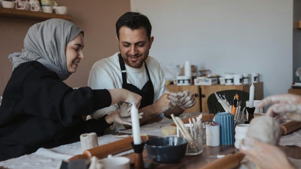 A smiling couple is sitting at a table in a bright room, working with clay or dough.