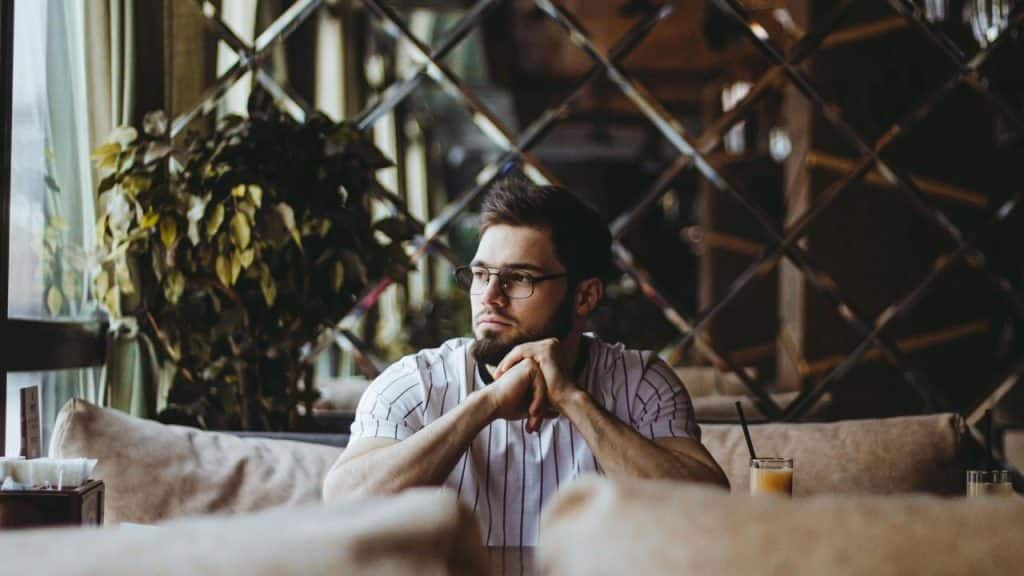 A man sitting in a cafe with his hands clasped, looking out the window thoughtfully.