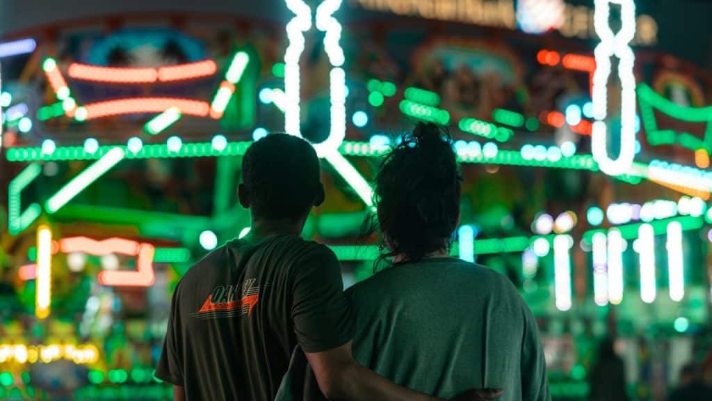A couple with their arms around each other's backs stands facing away from the camera, looking at a brightly lit carnival ride or fair attraction at night.
