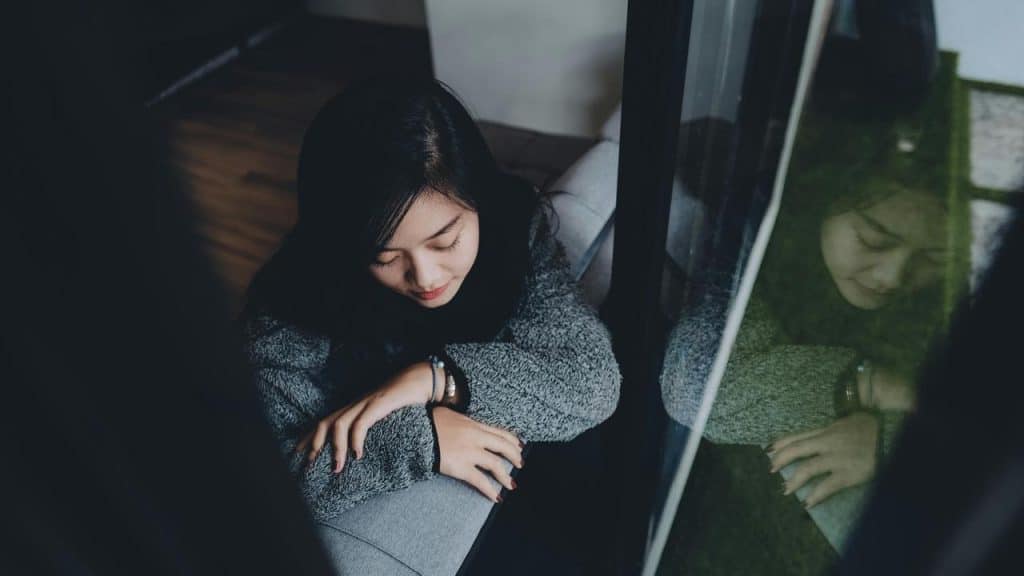 A woman sitting by a window with her head bowed and eyes closed.