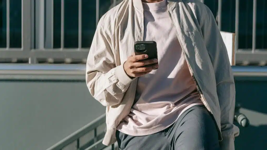 A person wearing a light jacket holding a smartphone while sitting outdoors.