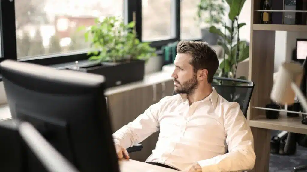 A man sitting at a desk in an office looking out the window thoughtfully.