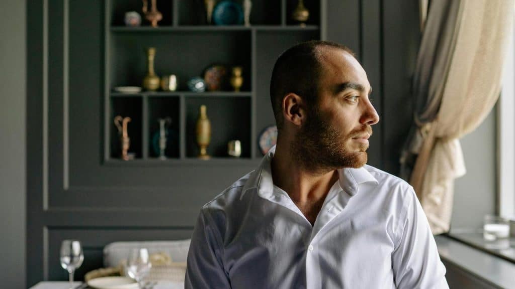 A man in a white shirt sitting indoors and looking out the window thoughtfully.
