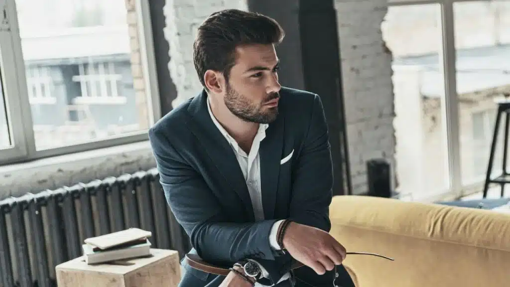 A well-groomed man in a dark suit jacket sitting and looking away in a bright, modern room.