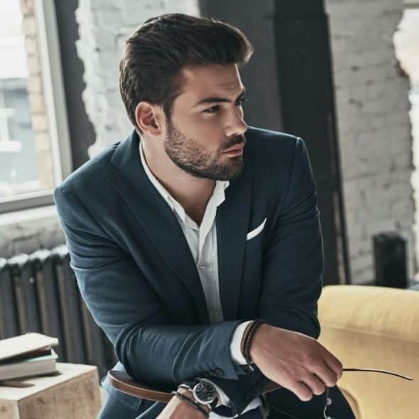 A well-groomed man in a dark suit jacket sitting and looking away in a bright, modern room.