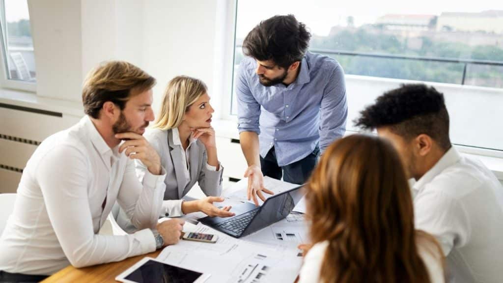 A diverse group of professionals discussing documents around a laptop on a table.