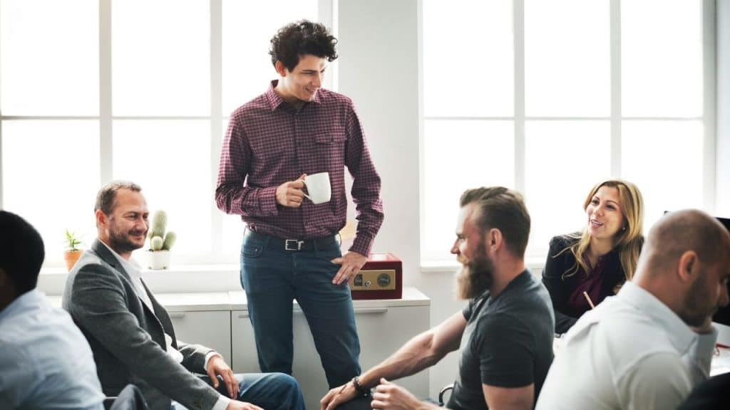 A smiling man stands holding a mug in a casual meeting with seated colleagues.