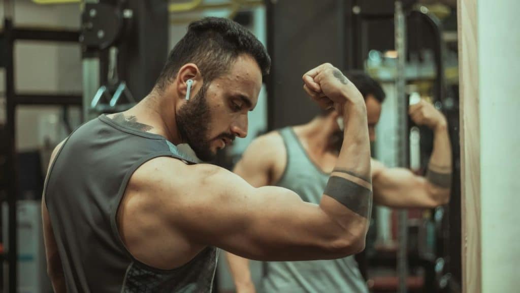 A bearded man in a tank top flexing his bicep while looking at his reflection.