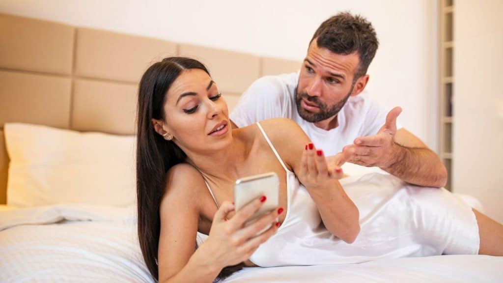 A concerned man gestures while a woman checks her phone lying on a bed.