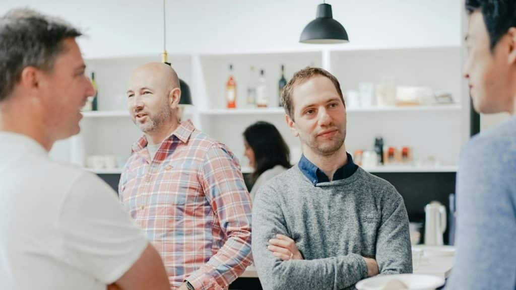 Three men talking and smiling while standing in a brightly lit kitchen or breakroom.