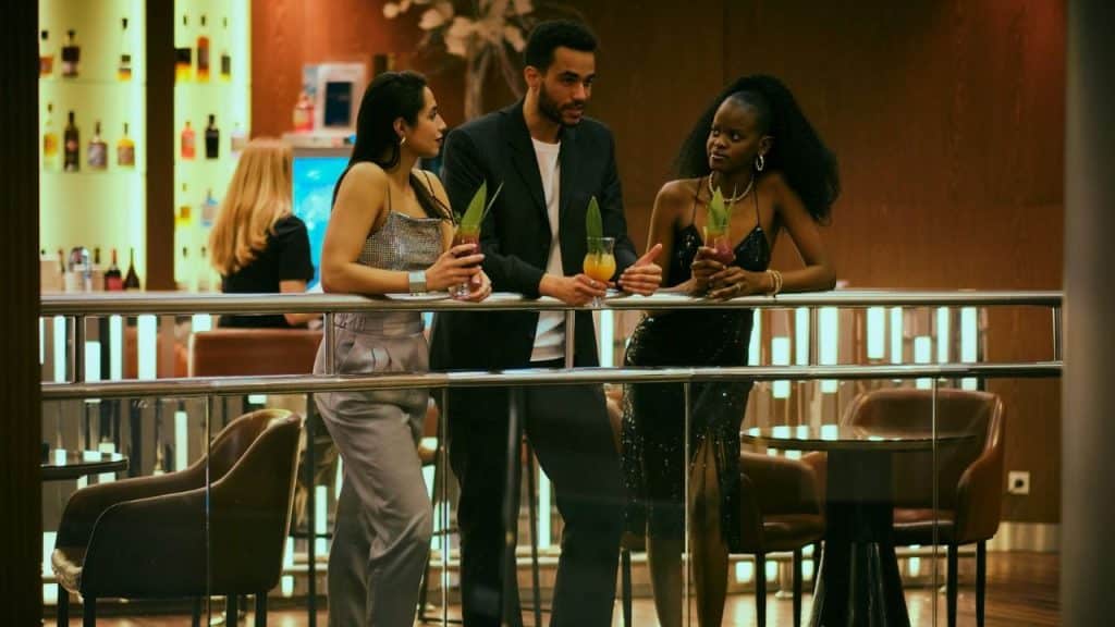 Three people dressed formally, holding cocktails, talking at a bar railing.