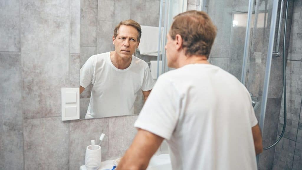 A serious man in a white t-shirt looks at his reflection in a bathroom mirror.