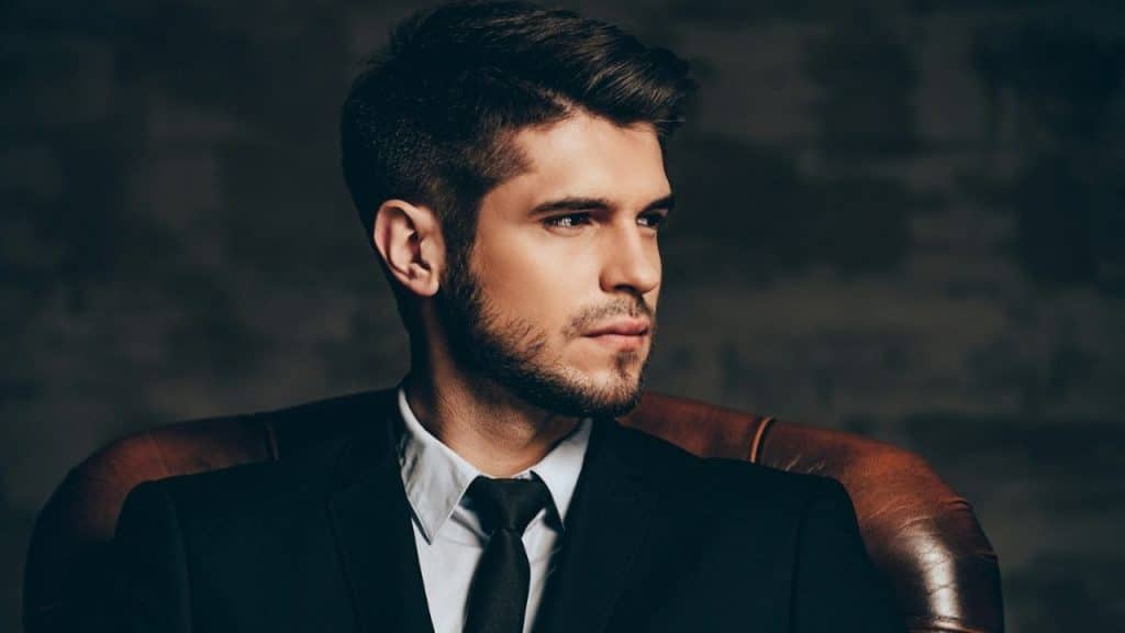 Young man in a black suit and tie sitting in a leather chair.