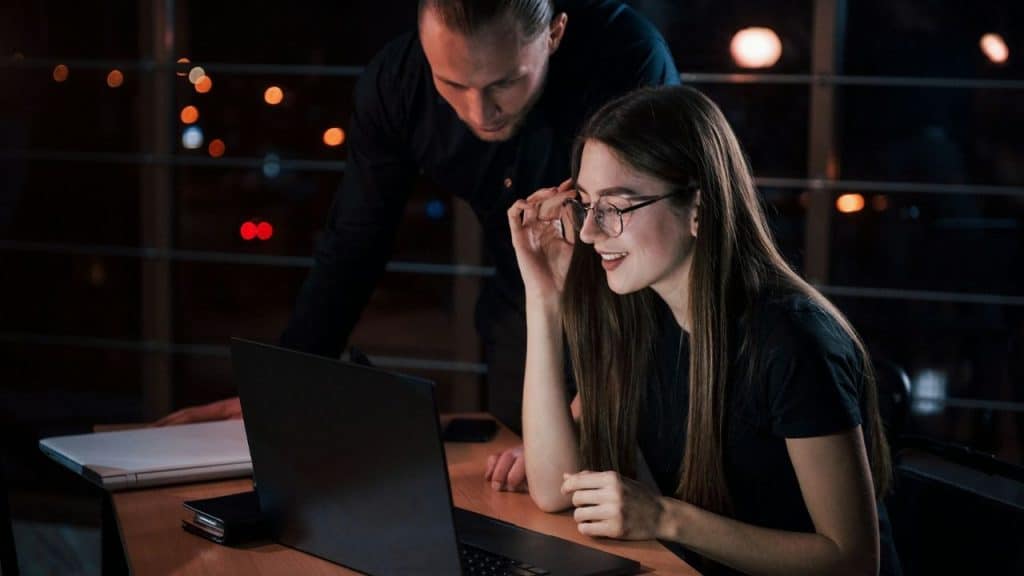 Two people working on a laptop at a desk in a dark office at night.