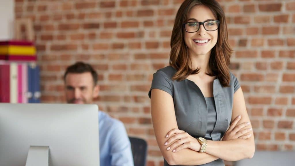 A smiling woman wearing a gray dress and glasses stands with arms crossed in an office.