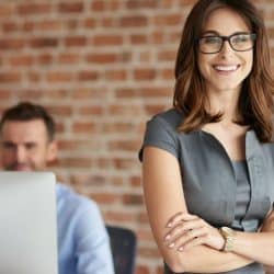 A smiling woman wearing a gray dress and glasses stands with arms crossed in an office.