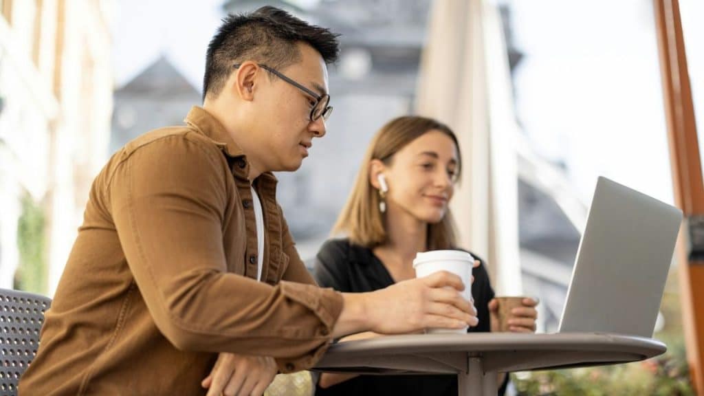 Two people sitting outdoors looking at a laptop, one holding a takeaway coffee cup.