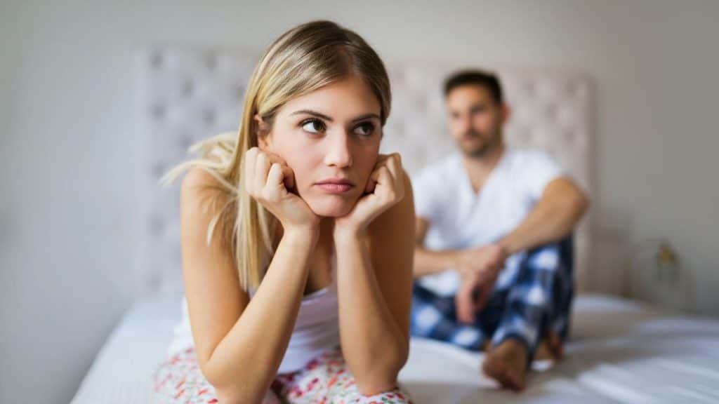 A sad woman sitting on a bed looking away, with a man out of focus behind her.
