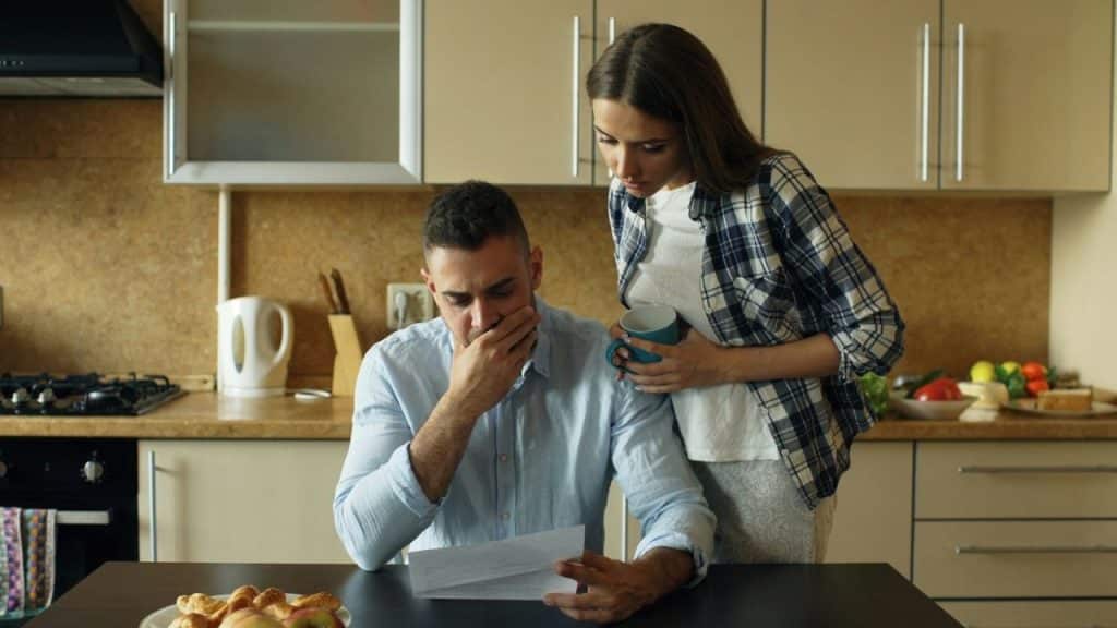 A worried man reading a document at a kitchen table while a woman looks over his shoulder.