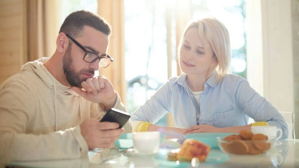 A woman smiling at a man who is focused on his phone during breakfast.
