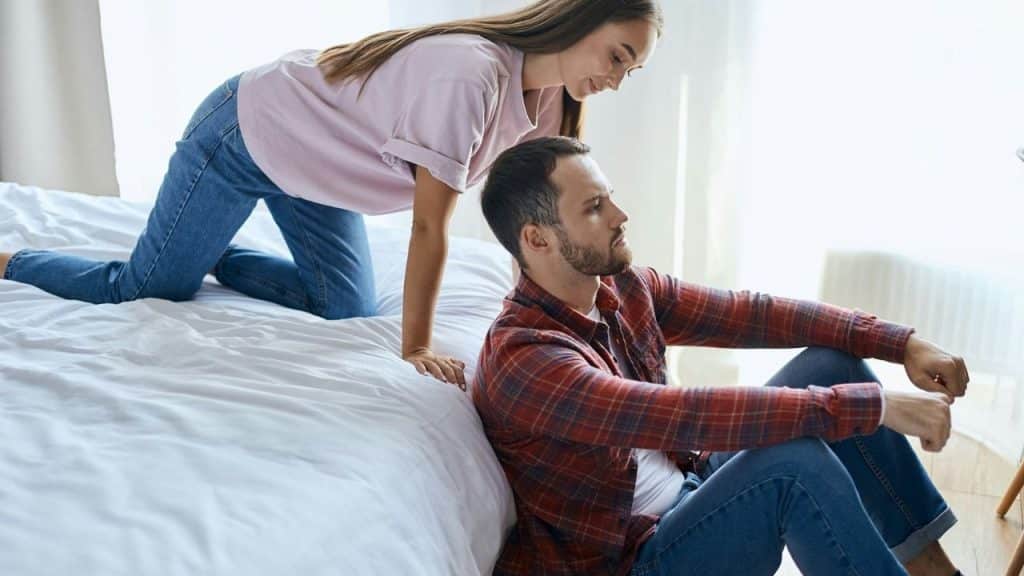 A woman on a bed looks down at a man sitting on the floor beside her.