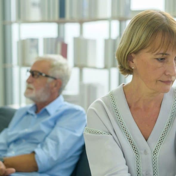 An older woman looks sad or distressed while an older man sits turned away from her on a couch behind her.