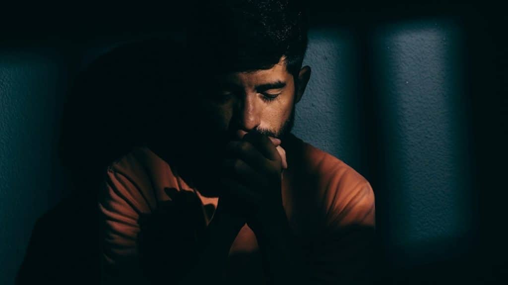 A bearded man with closed eyes holding clasped hands up to his mouth in a dark room.