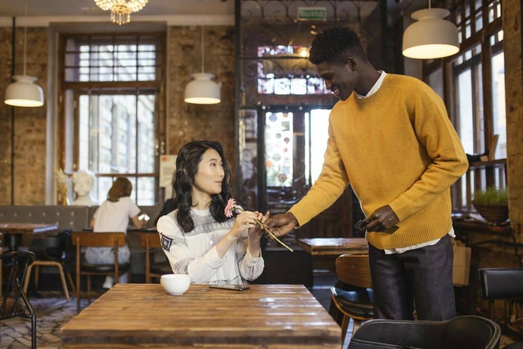 The man is giving the woman a pink flower in a café.
