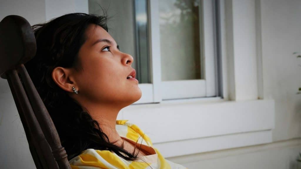 Woman sitting in a wooden chair, looking up and away from the camera, near a white window.