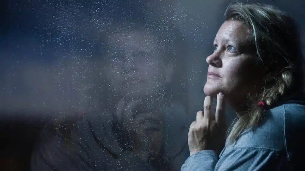 Woman in a grey shirt looking out a rain-streaked window, resting her chin on her hand.