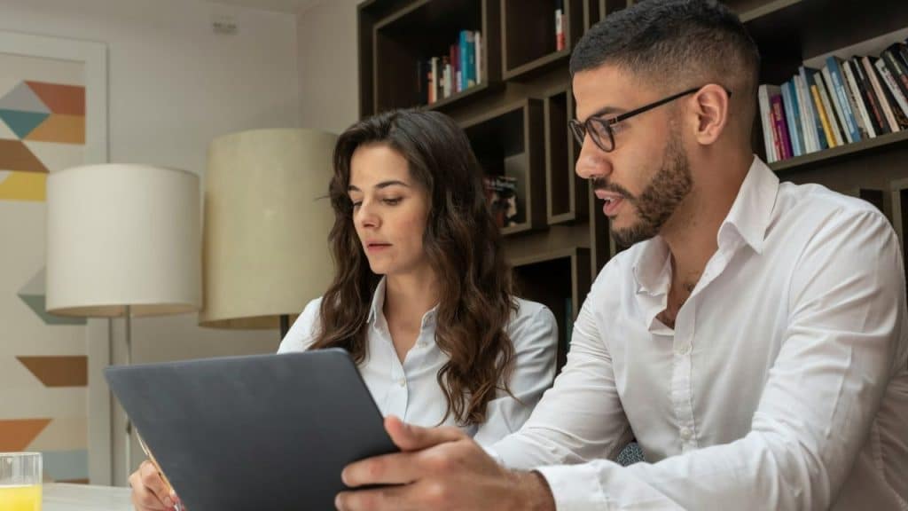 Two people in white shirts looking at a laptop together, with a bookcase in the background.
