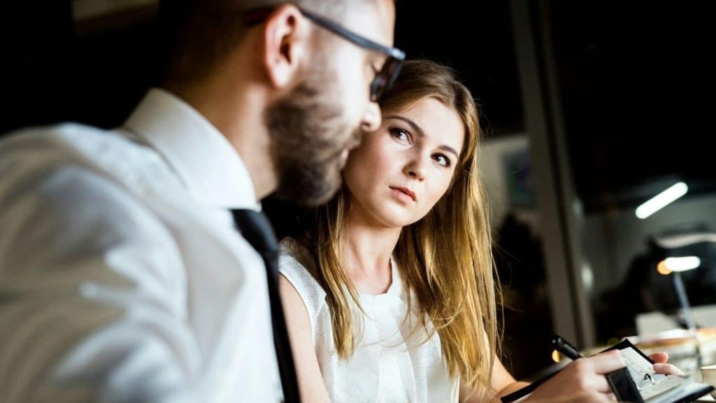 Woman holding a pen and notebook, looking intently at a man in a white shirt and tie.