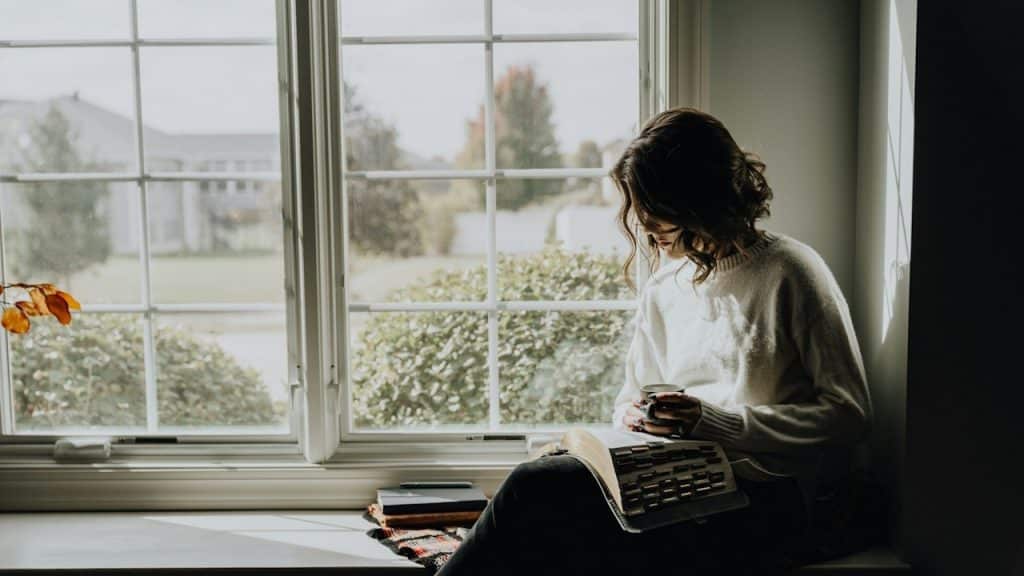 A woman sits on a sunlit window seat, reading a book and holding a mug, with a leafy neighborhood outside.