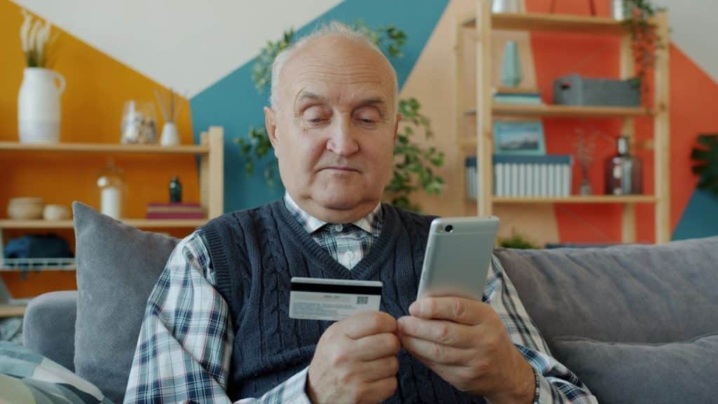 An elderly man holding a credit card and smartphone for online shopping.
