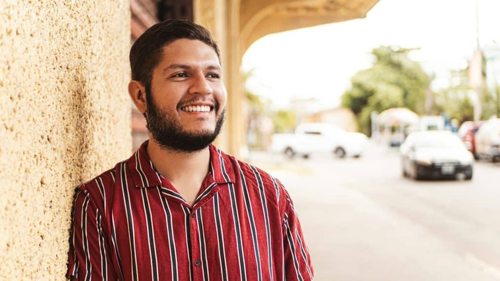 A smiling bearded man wearing a striped black and red shirt and standing in the street.