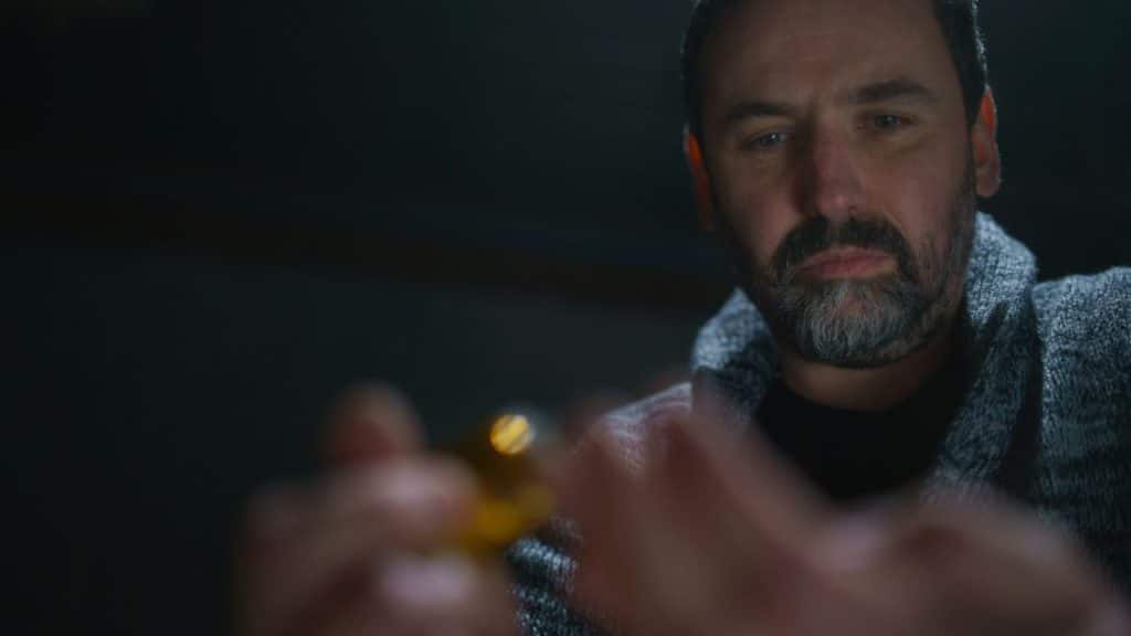 Close-up of a bearded man in a gray sweater holding a small, glowing object in the dark.