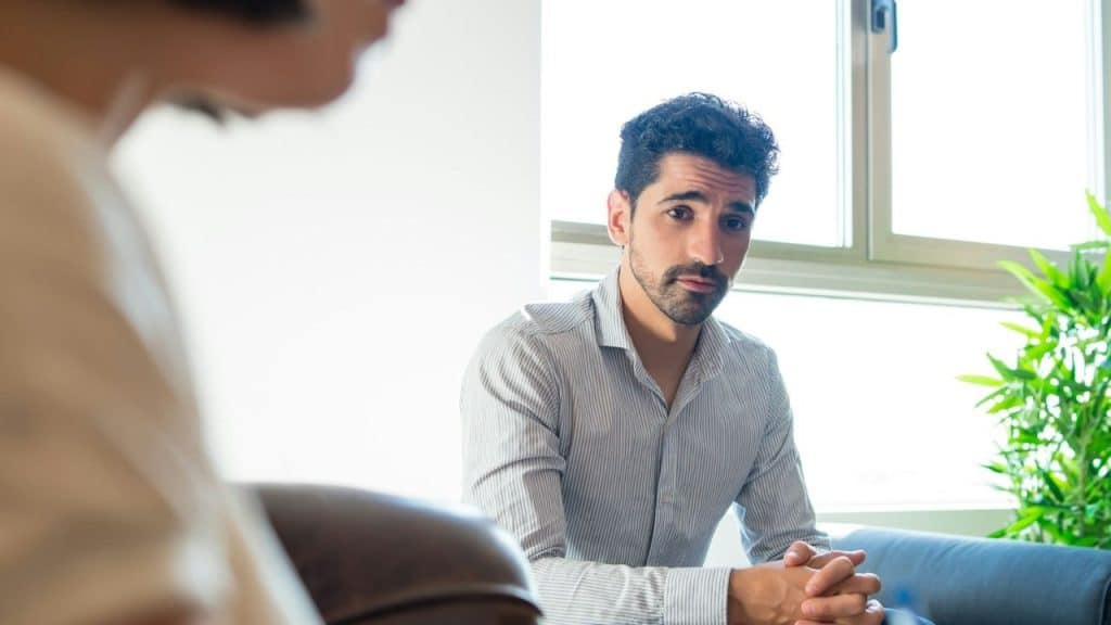 Bearded man in a striped shirt sitting, looking directly forward in a bright room.