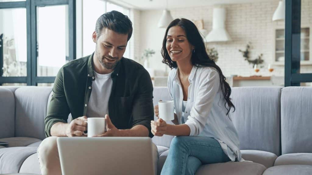 Smiling couple on a sofa holding mugs and looking at a laptop in a bright living room.