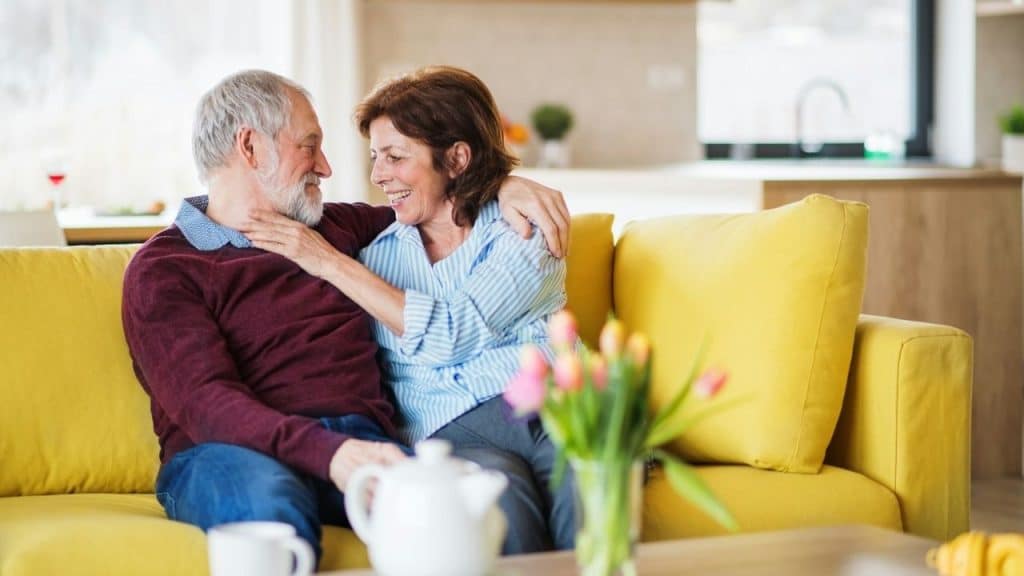 Smiling older couple hugging on a yellow sofa in a bright living room.