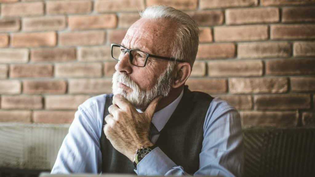 Older man with white beard and glasses contemplating with his hand on his chin.
