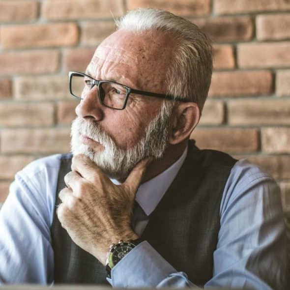 Older man with white beard and glasses contemplating with his hand on his chin.