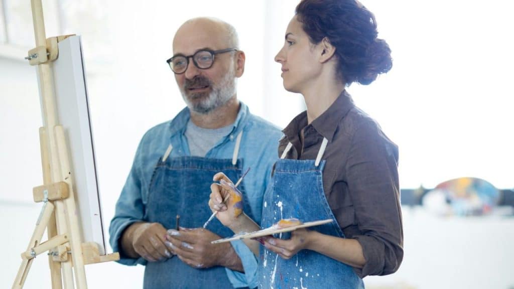 Two people in aprons, holding paint brushes and palettes, standing next to an easel.