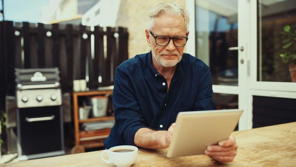 Older man in glasses reading a tablet outdoors with a coffee cup nearby.