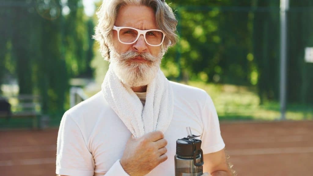Older man with white beard, white glasses, and a towel holds a water bottle outdoors.