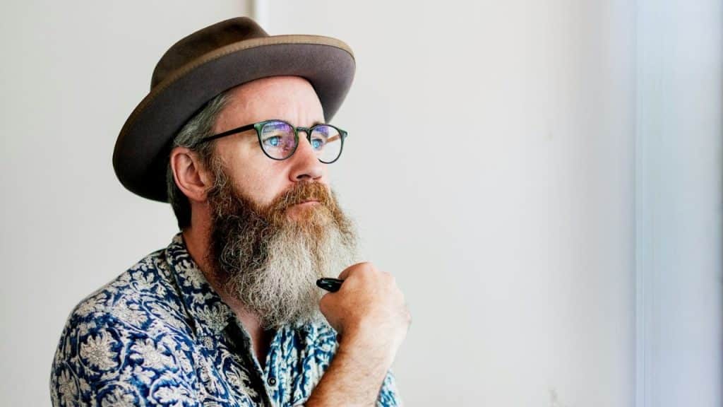 Man with a long gray beard, fedora, and glasses looking thoughtful indoors.