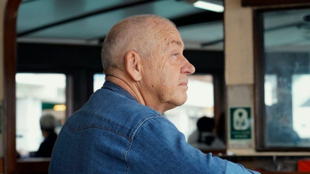 Side profile of an older, bald man in a denim shirt looking away indoors.