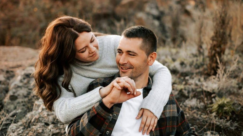 A couple sitting outdoors, the woman hugging the man as they smile at each other.