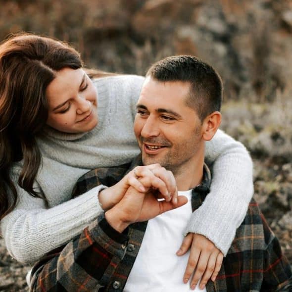 A couple sitting outdoors, the woman hugging the man as they smile at each other.