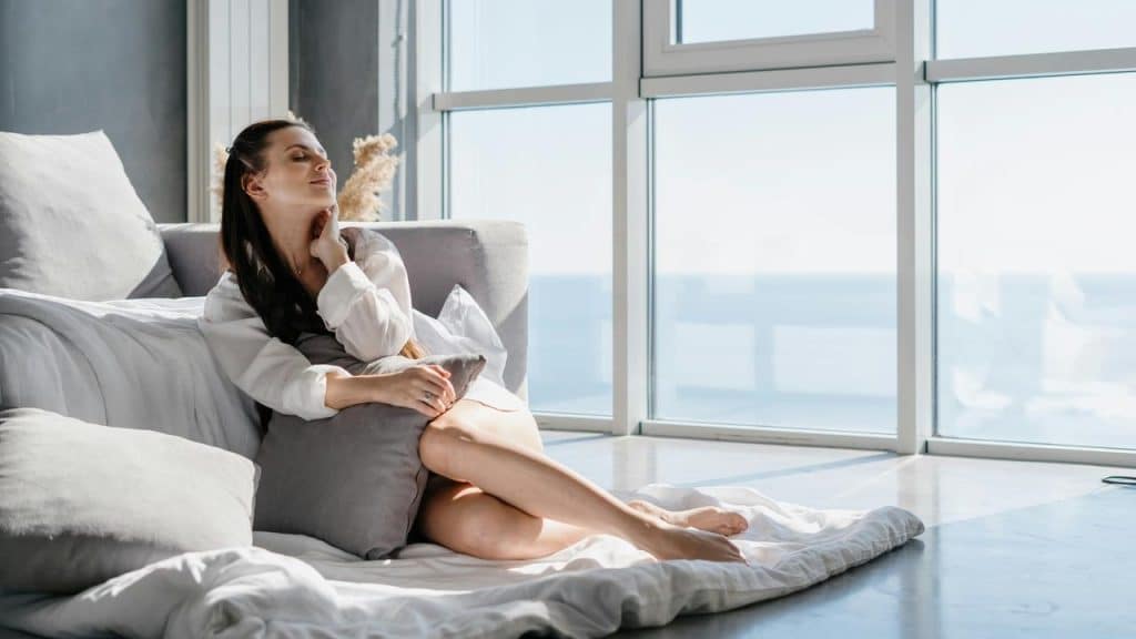 A woman relaxes on the floor by a large window, enjoying the sunlight.